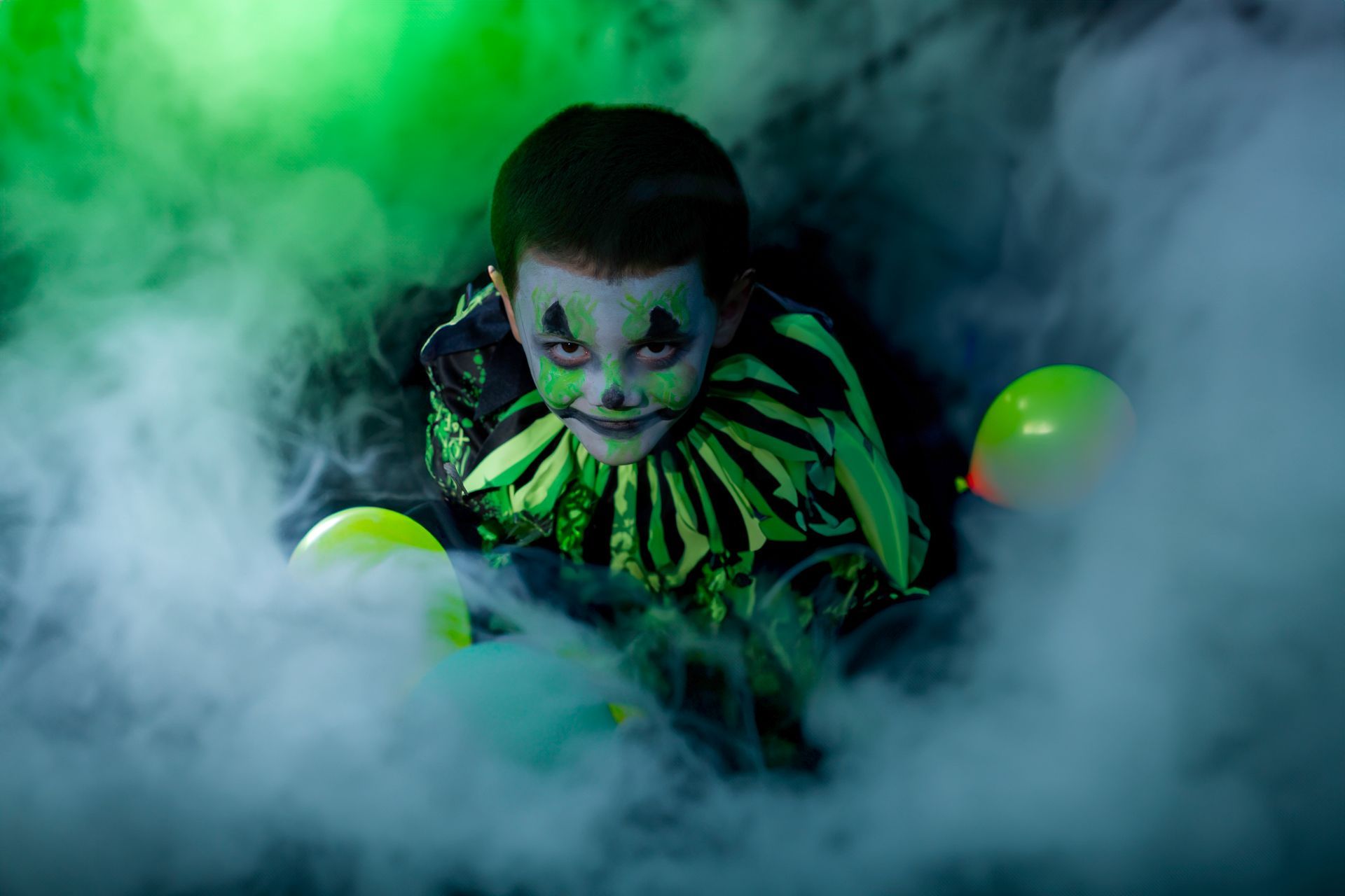 A young boy dressed as a clown is surrounded by balloons and smoke.