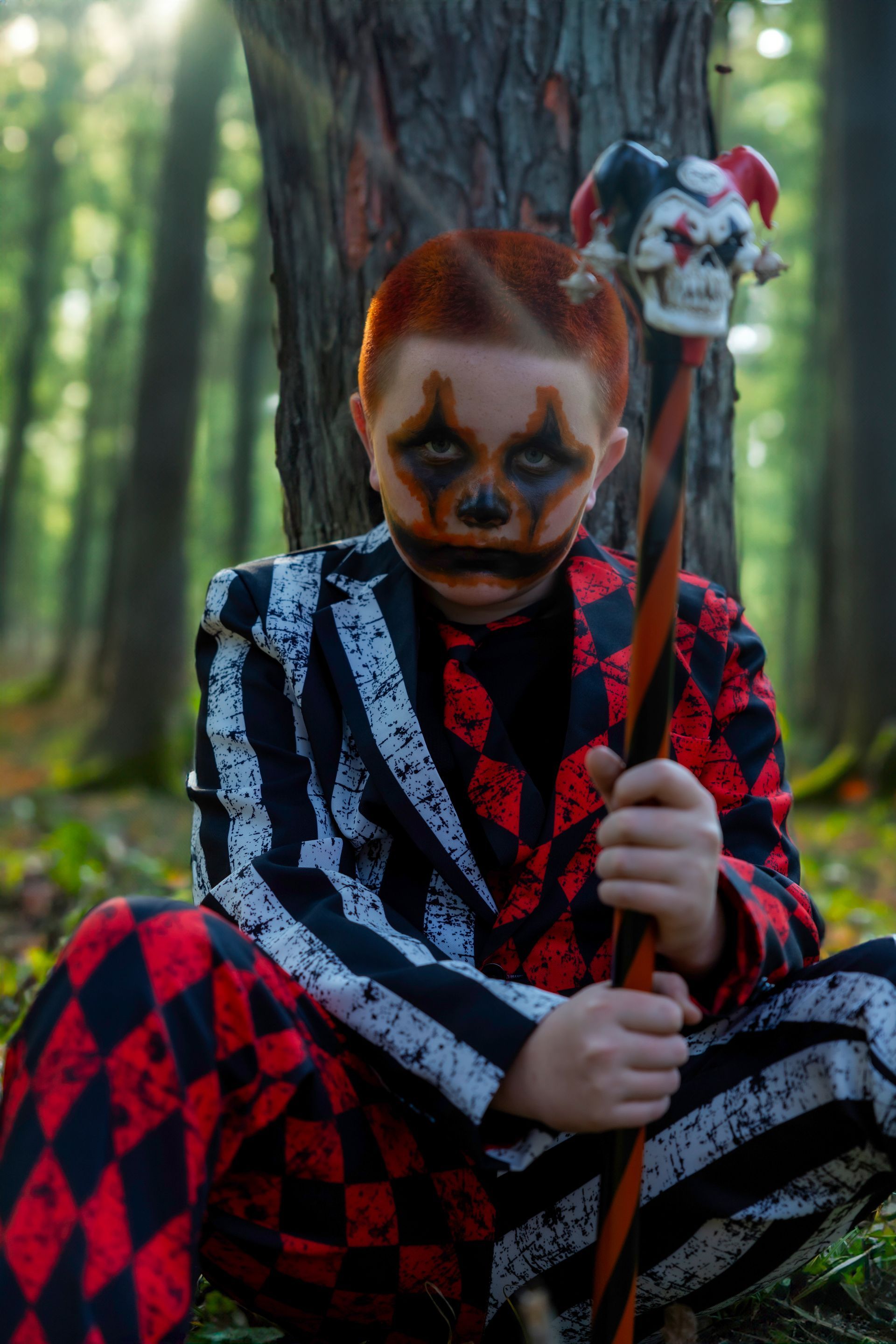 A young boy dressed as a clown is sitting in the woods holding a cane.