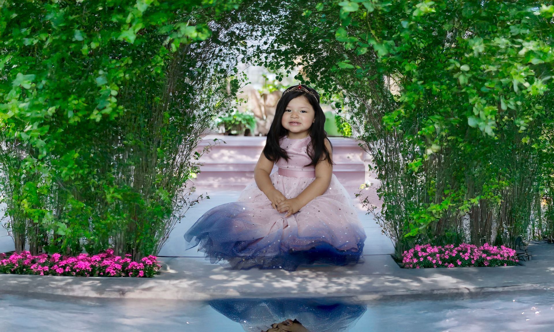 Young girl in pink and blue dress, smiling in garden, framed by foliage.