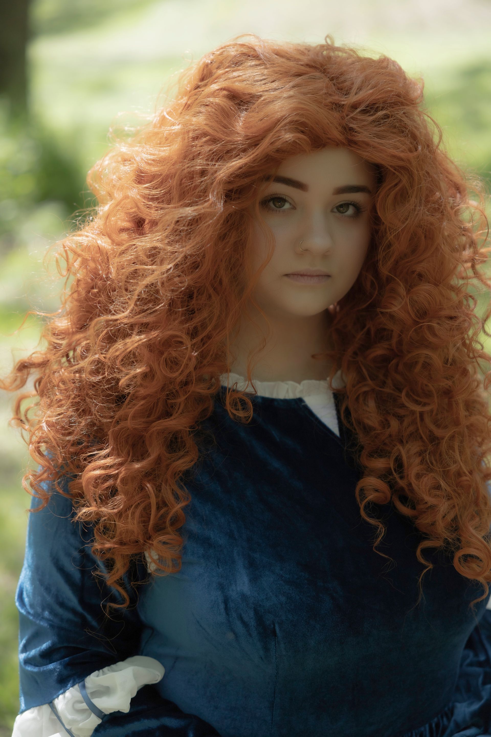 Woman with voluminous red curly hair in a blue dress, looking forward with a neutral expression, outdoors.