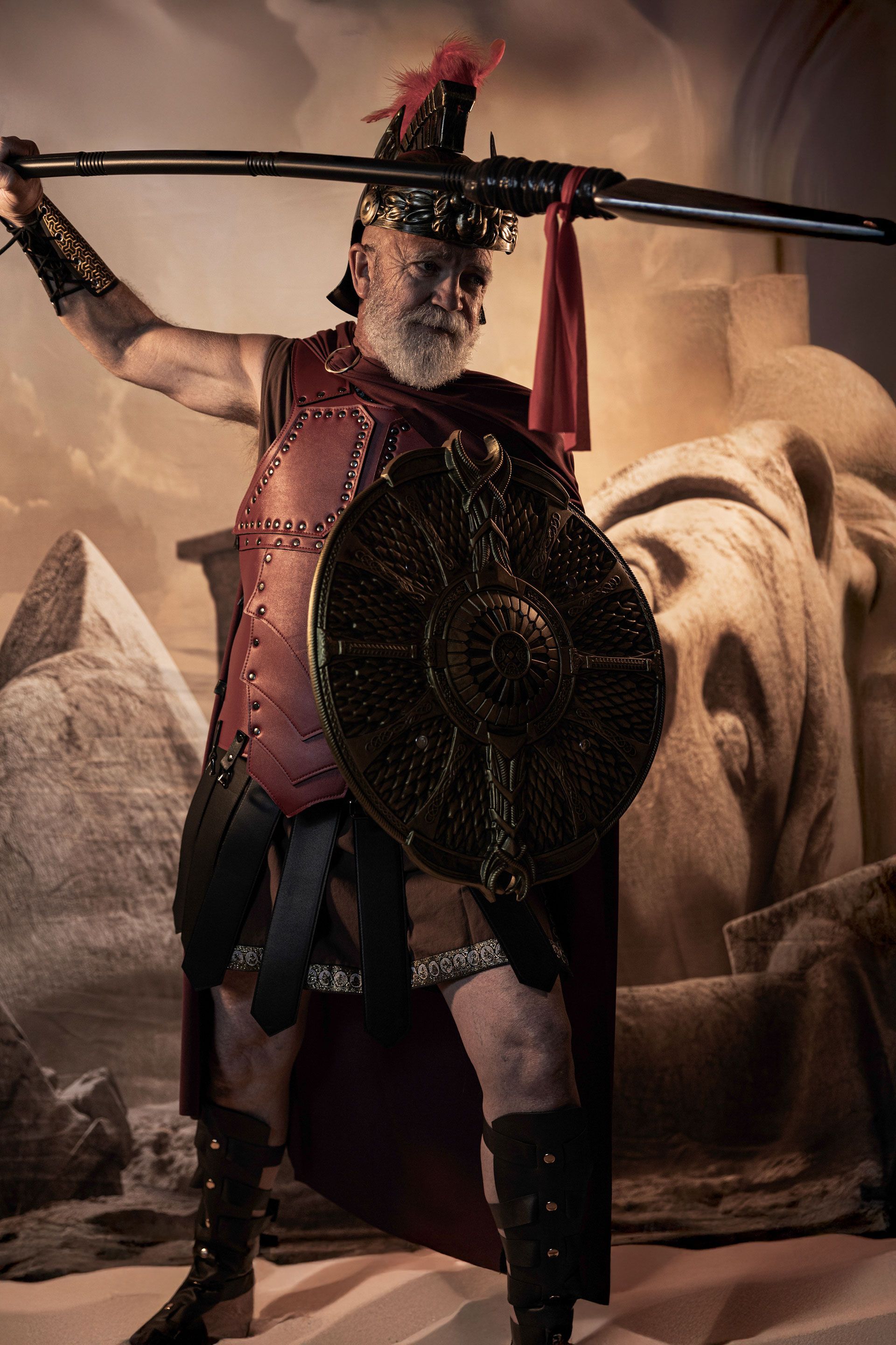 Man in Roman armor, holding spear and shield, posed against a backdrop of desert and ruins.