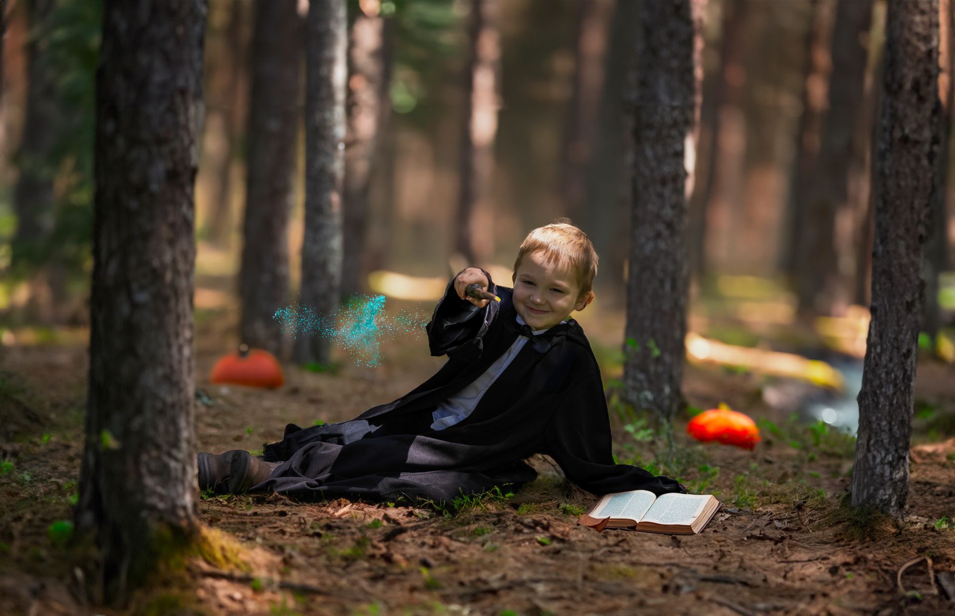 A little boy dressed as a wizard is sitting in the woods with a book and a wand.
