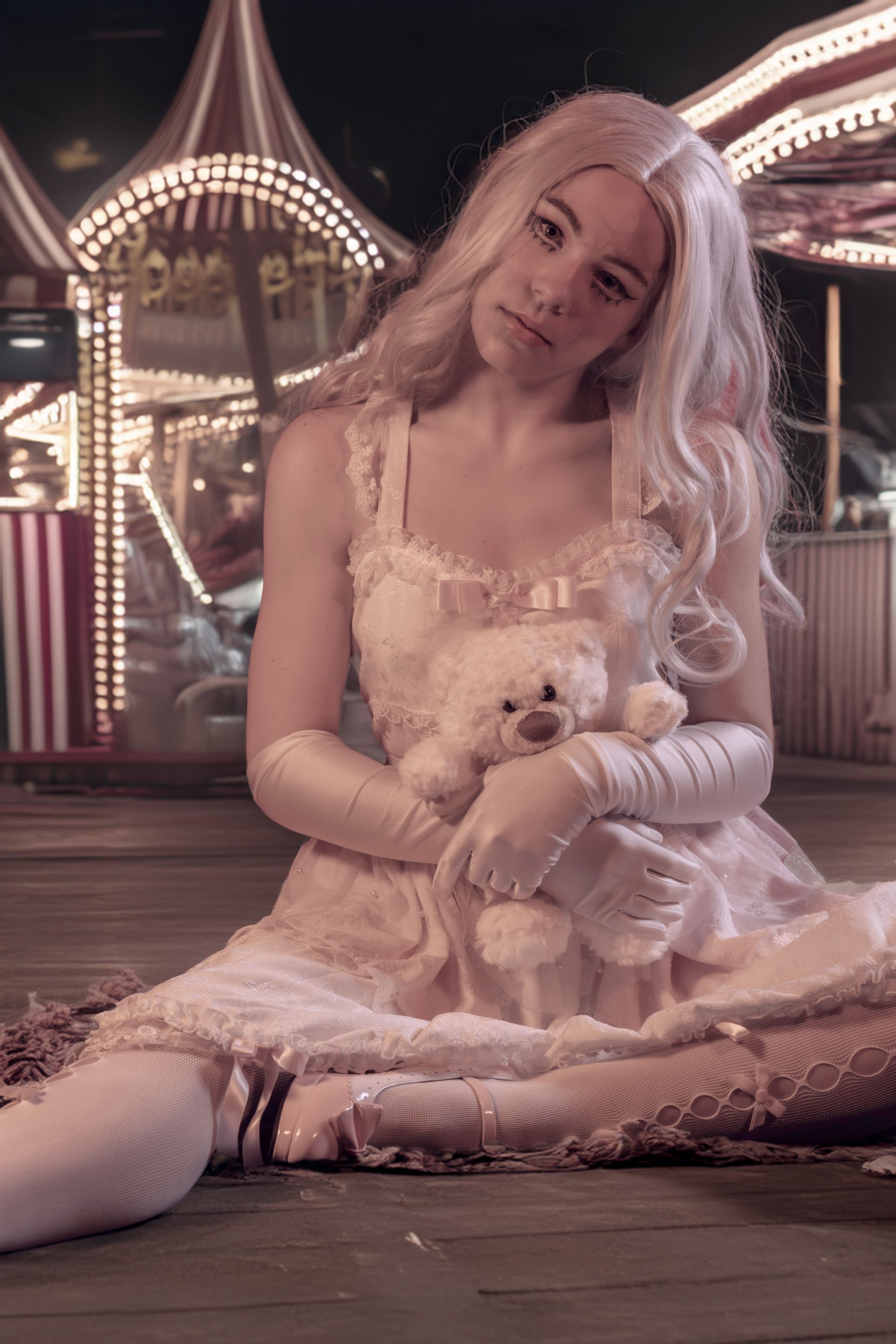 Woman in a pink dress and gloves holds a teddy bear at a carnival.
