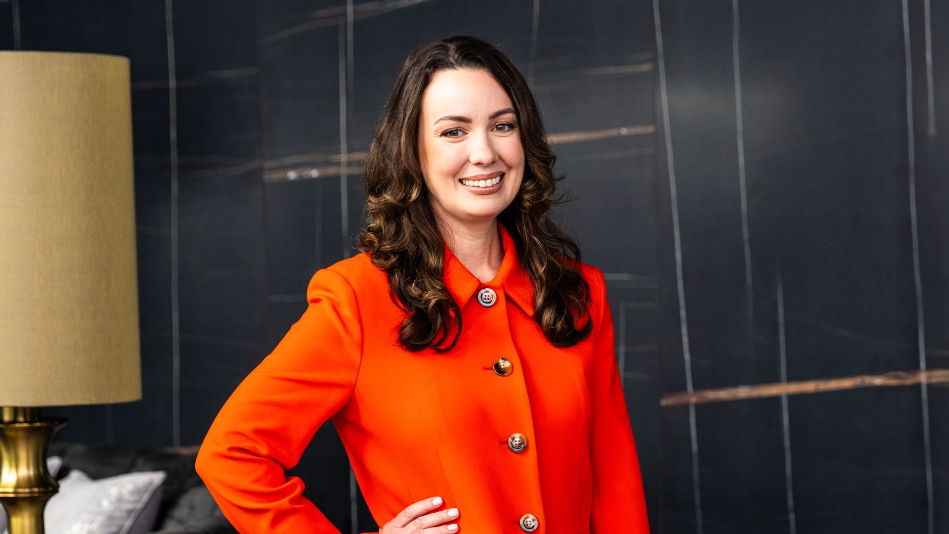 A woman in an orange coat is standing in front of a lamp.