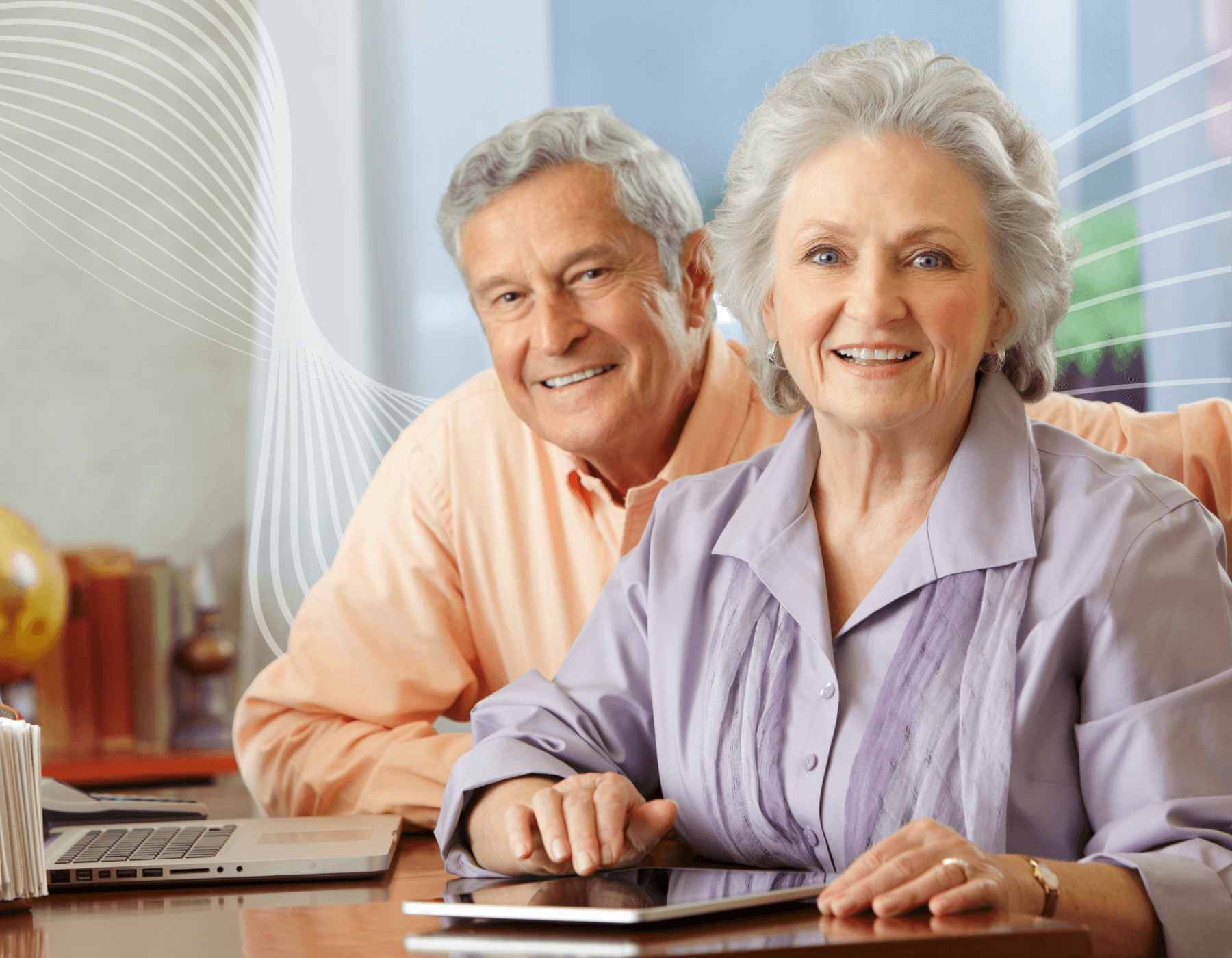 An elderly couple is sitting at a table with a laptop and a tablet.