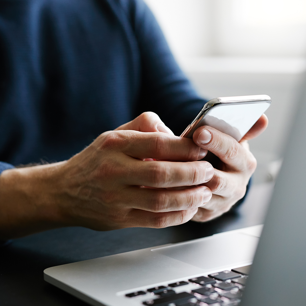 A man is holding a cell phone in his hands in front of a laptop.