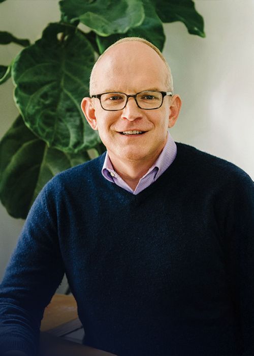 A man wearing glasses and a blue sweater is sitting in front of a plant.