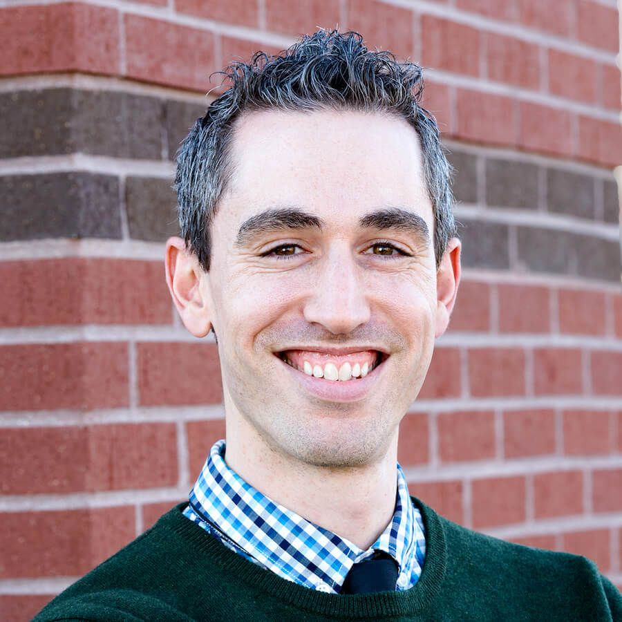 A man wearing a green sweater and tie is smiling in front of a brick wall.