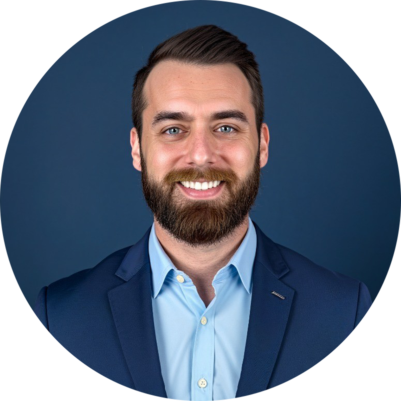 Ryan Maggi with a brown beard smiling, wearing blue suit and button-down shirt, set against a dark blue background.
