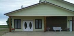 Small beige building with white double doors, brick accents, and a bench on the porch.