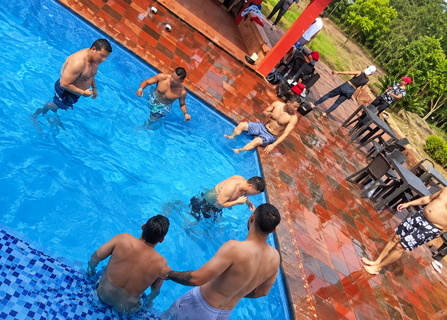 Personas chapoteando en una piscina azul, junto a una terraza de baldosas rojas y unas tumbonas.