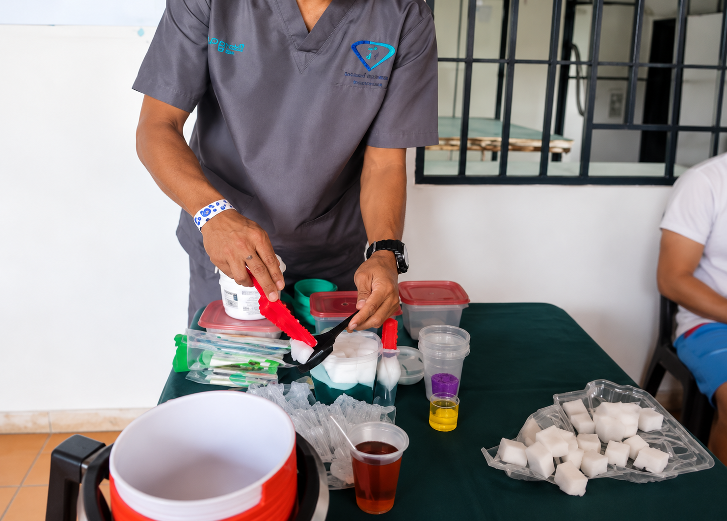 Una persona vierte líquido en vasos sobre una mesa con material de laboratorio y muestras blancas al aire libre