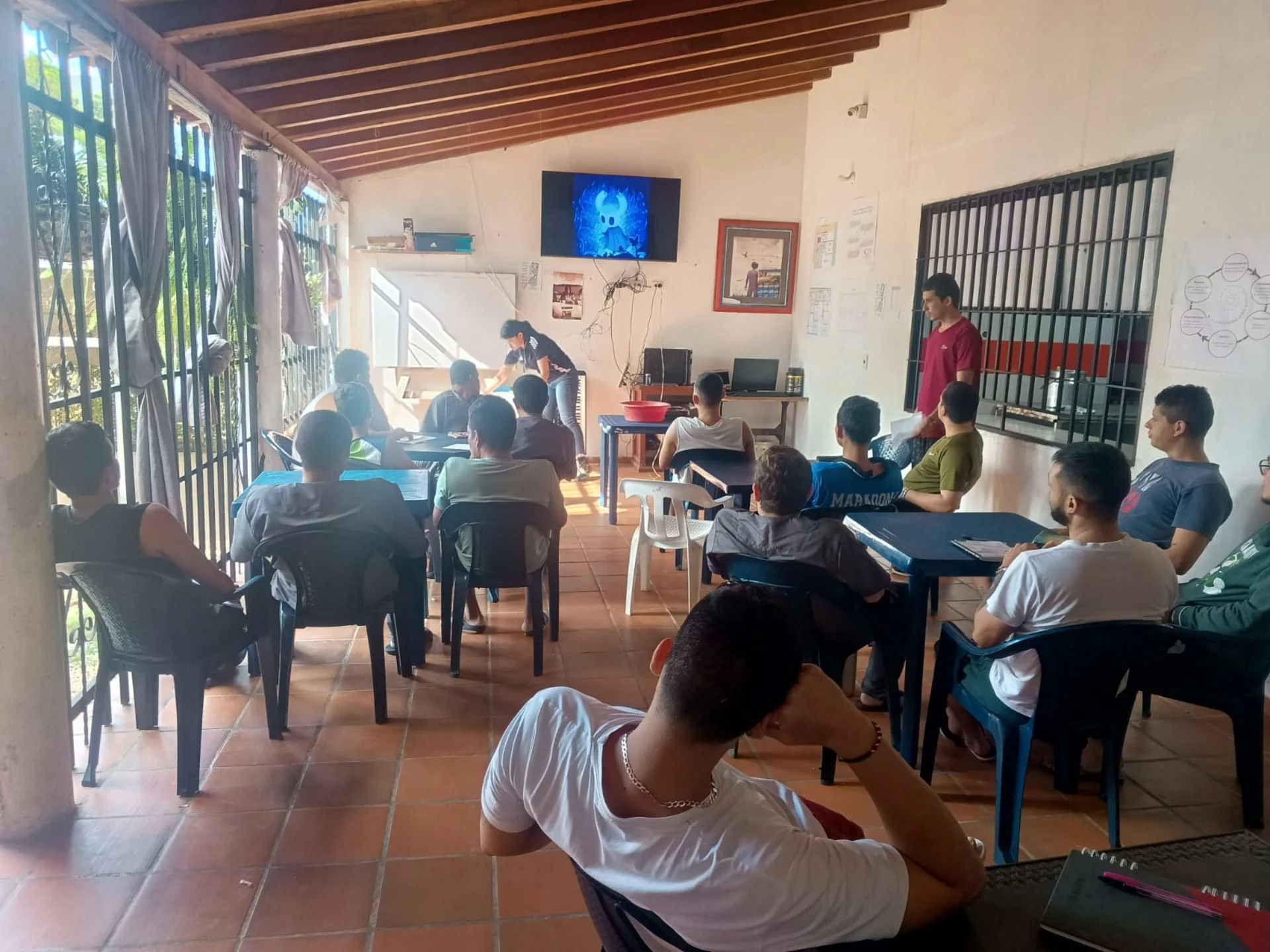 Personas sentadas en un aula viendo una lección en televisión en un patio cubierto