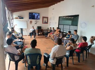 Personas sentadas en círculo en una pequeña sala de reuniones con una pantalla de televisión y una pizarra, conversando juntas