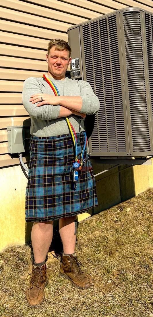 Person standing outdoors in a blue plaid kilt, gray sweater, and brown boots beside stacked equipment.