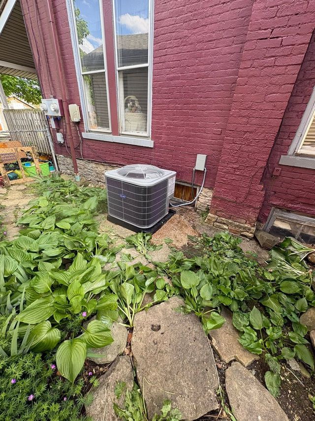 Outdoor AC unit beside a brick house, surrounded by overgrown green plants and cracked stone path.