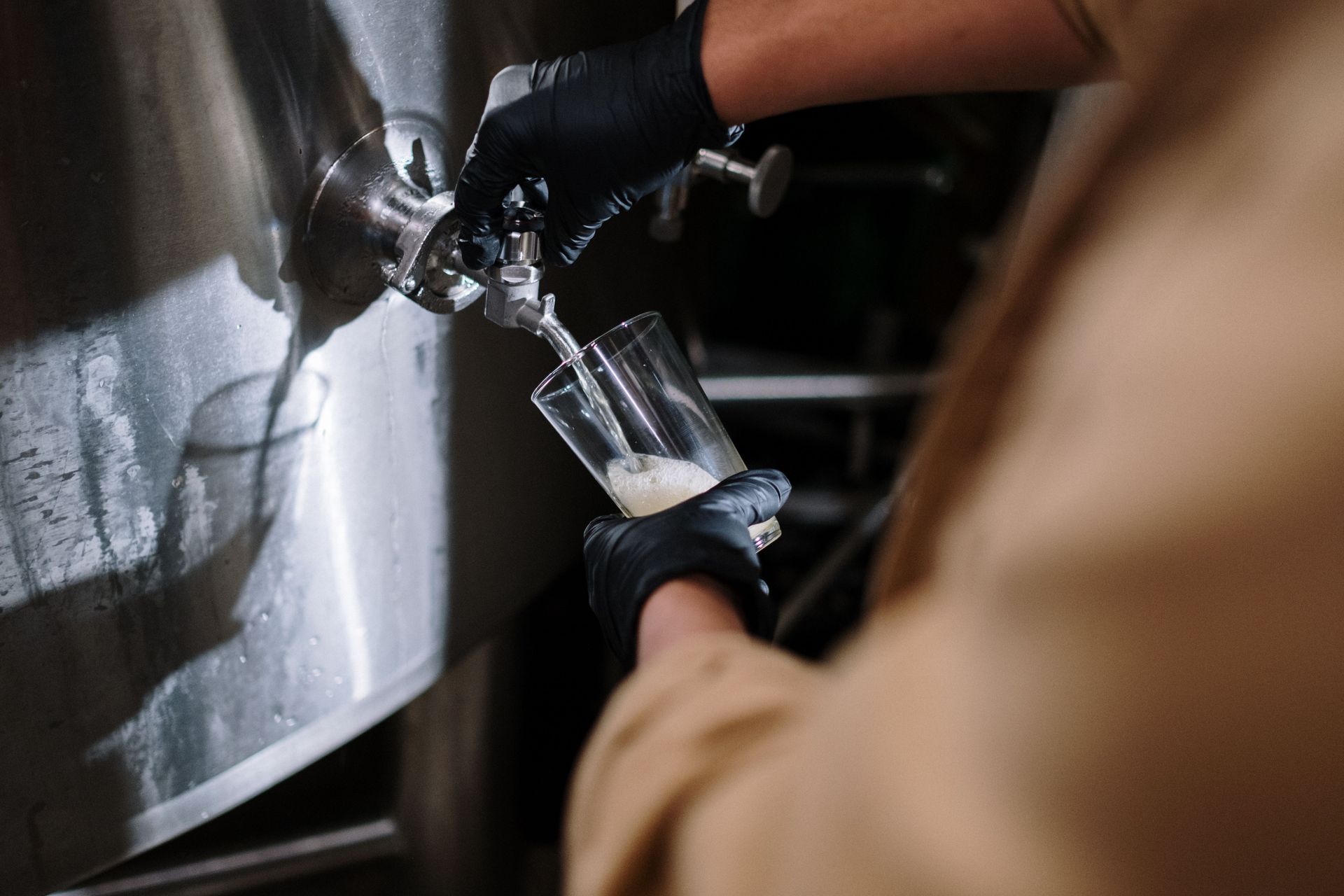 Gloved hands pouring liquid into a glass under a metal tap in a dimly lit kitchen or bar setup