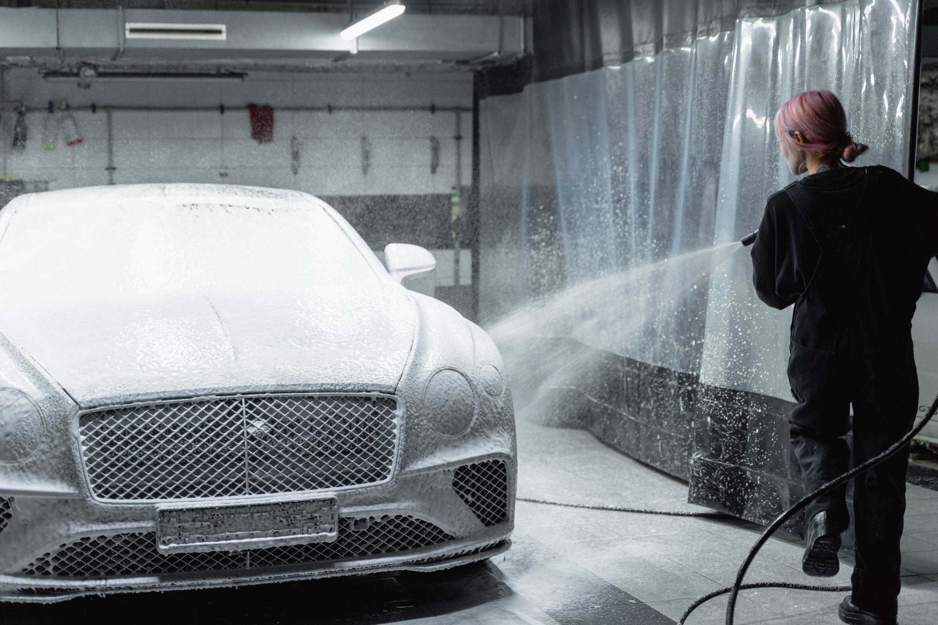 Person pressure-washes a white car covered in foam inside a garage.