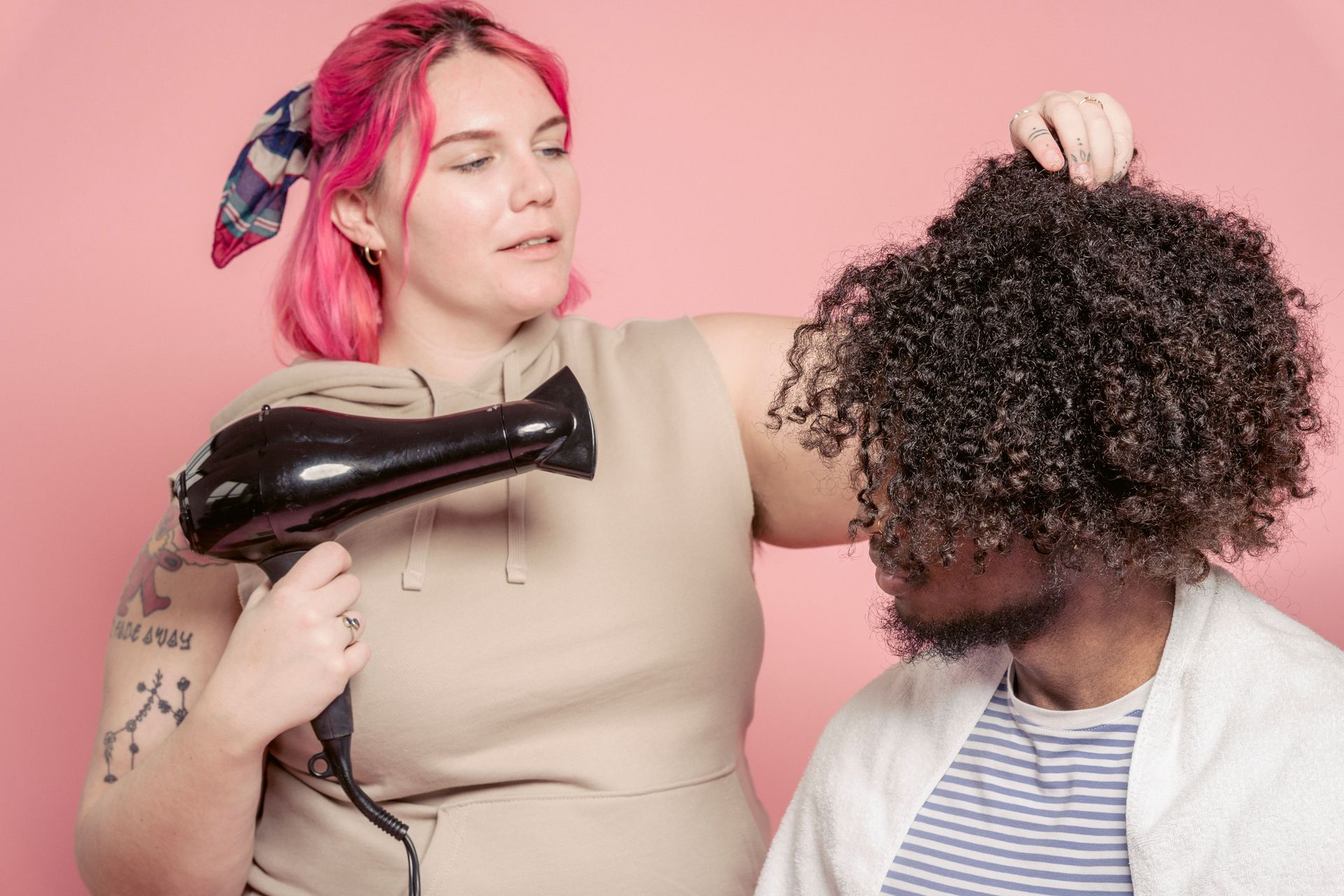 Woman blow-drying another person's curly hair against a pink background