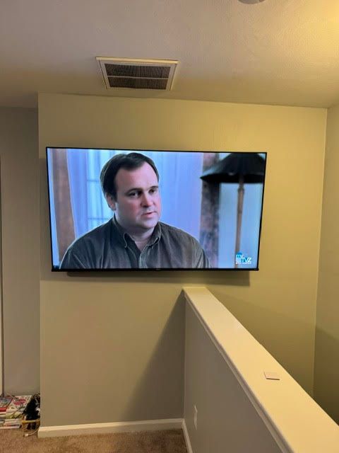 TV mounted on a beige wall showing a man in a gray shirt, viewed from a stair landing