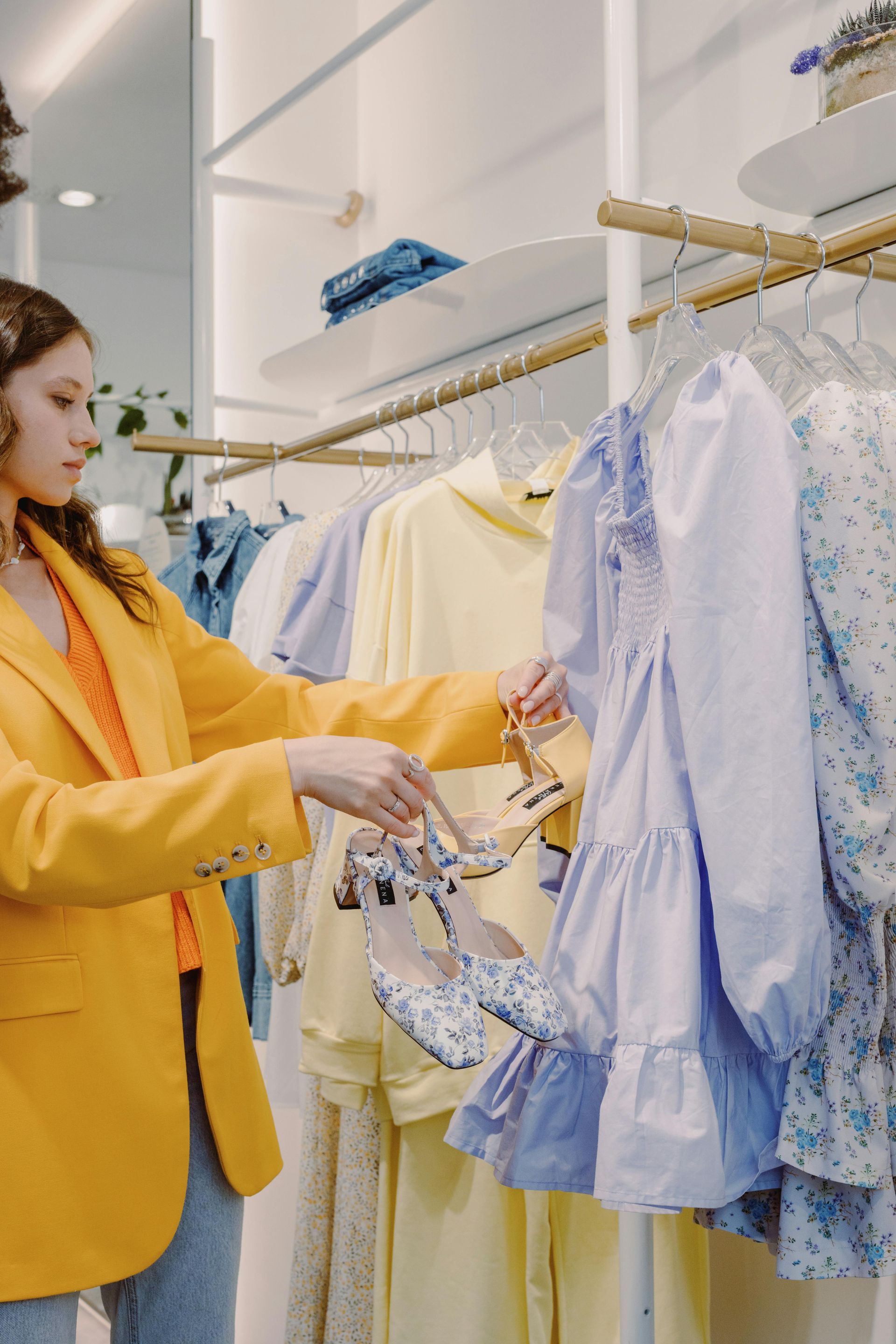 Woman in yellow jacket holding shoes, looking at a light blue dress in a clothing store.