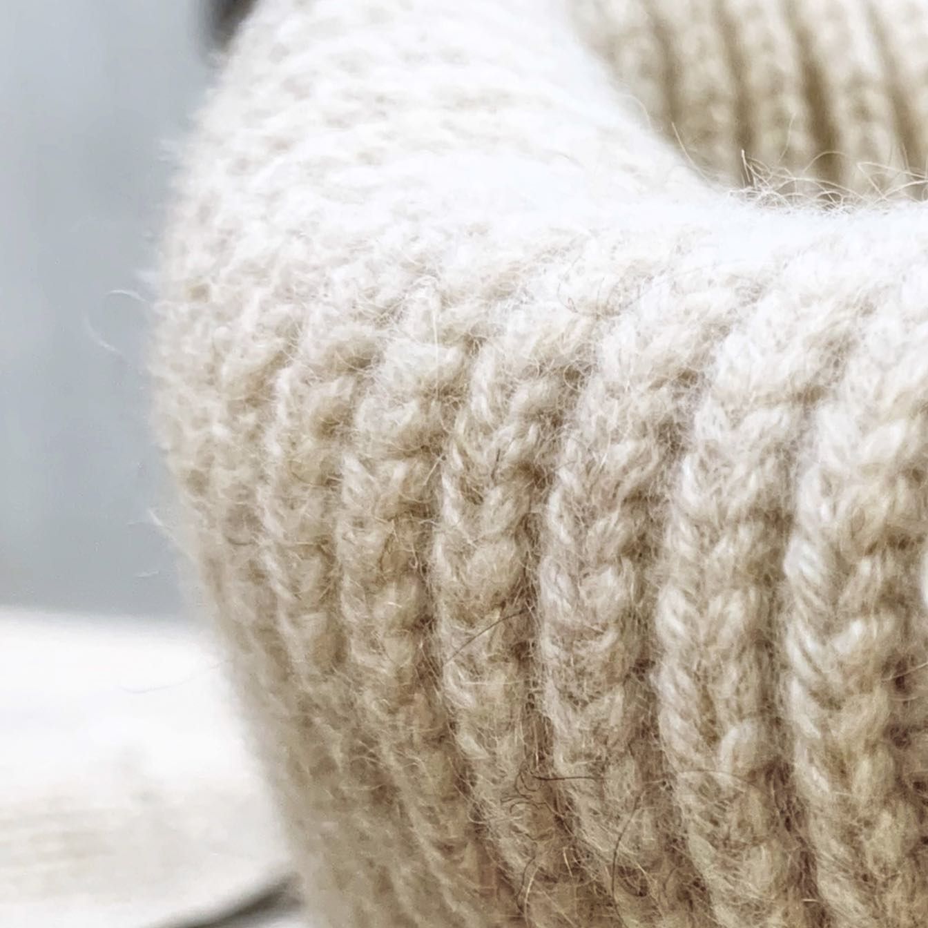 Primo piano della manica di un maglione di lana color crema su uno sfondo grigio tenue.
