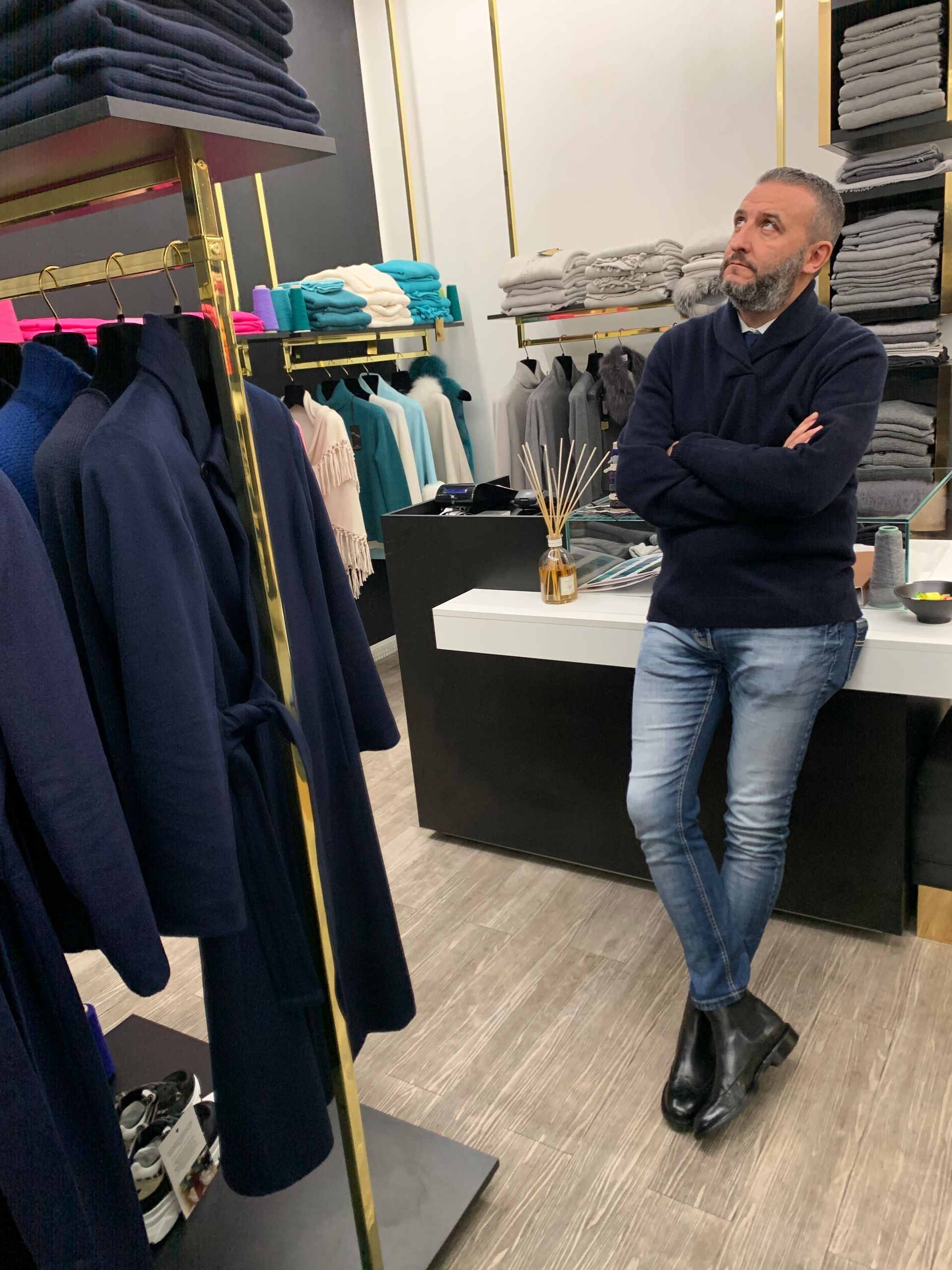 Un uomo è in piedi in un negozio di abbigliamento, con le braccia incrociate, accanto agli scaffali di giacche e camicie.