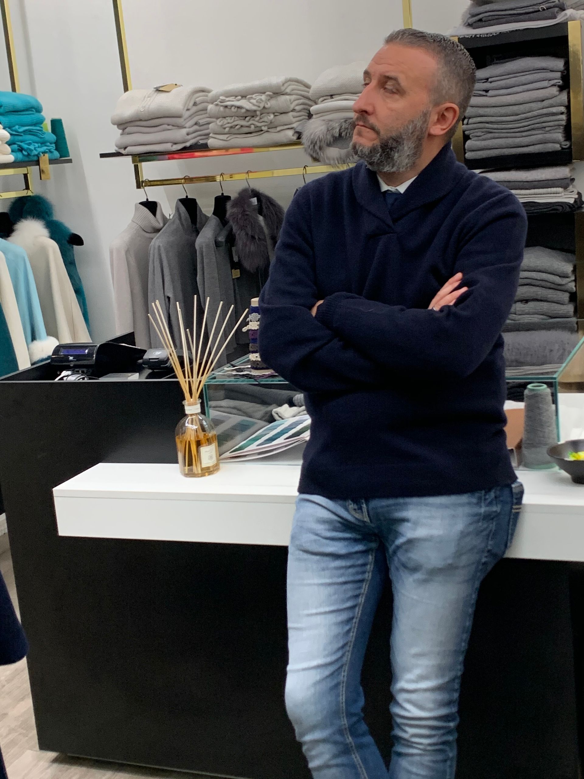 Un uomo in piedi con le braccia incrociate in un negozio di abbigliamento, accanto ad asciugamani piegati e articoli in esposizione.