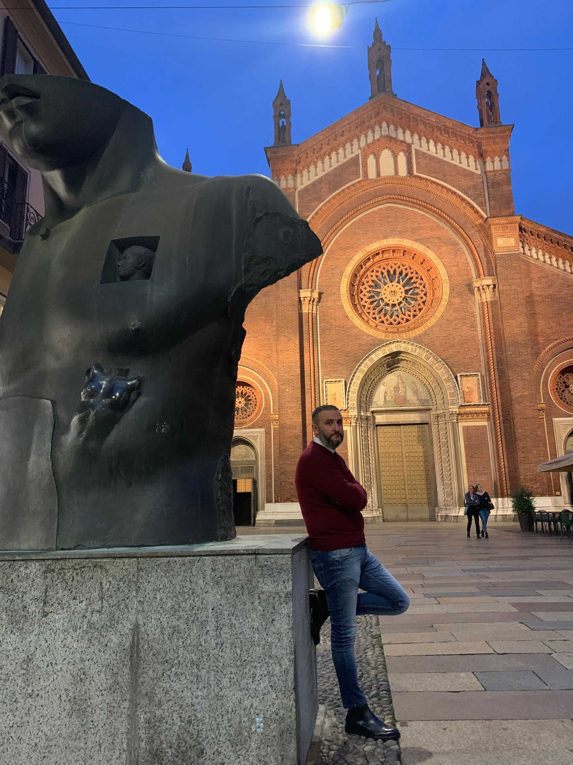 Un uomo in piedi accanto a una statua scura in una piazza al crepuscolo, con una cattedrale di mattoni illuminata sullo sfondo.