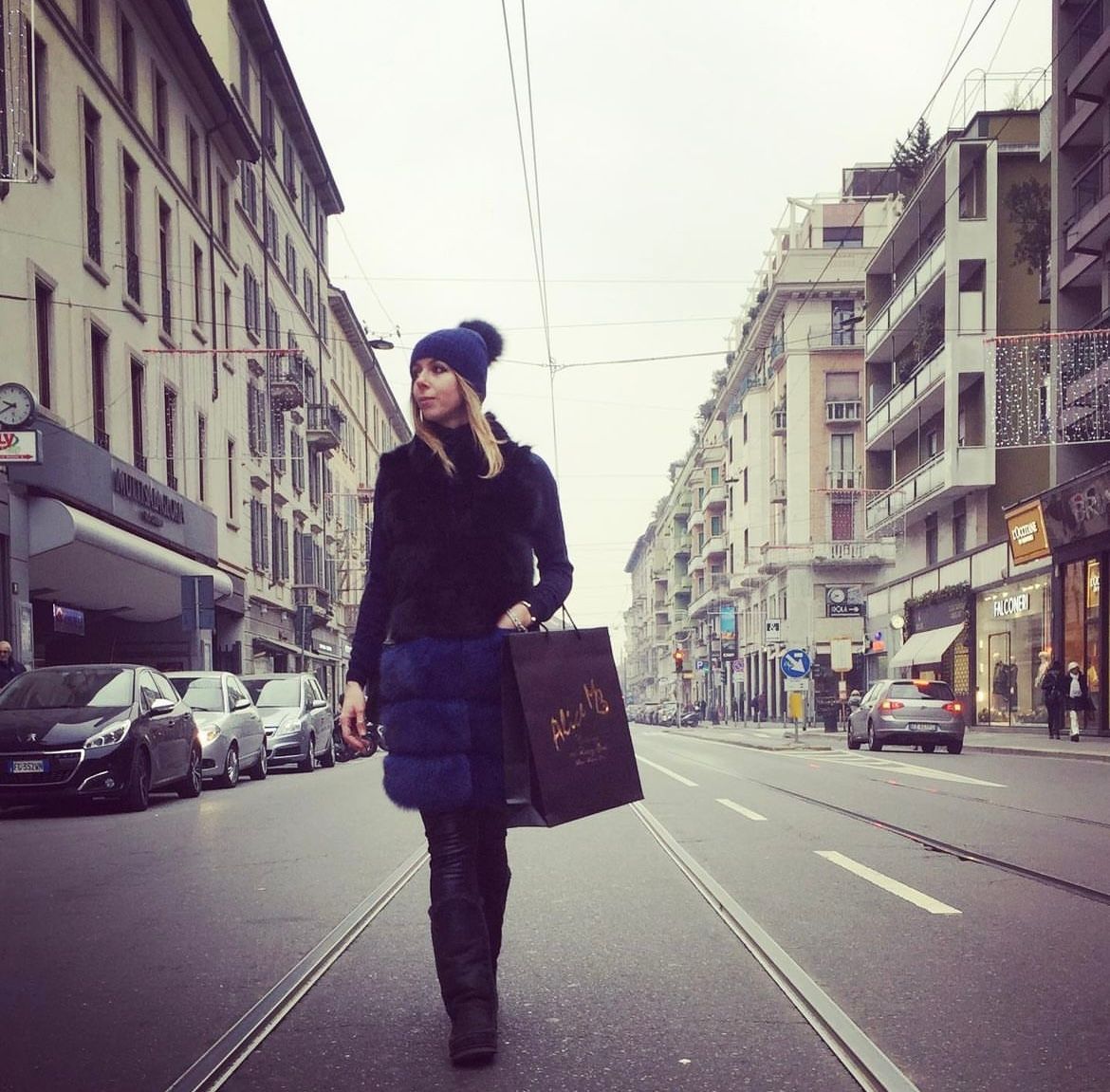 Una donna cammina per una strada cittadina, indossando un cappello blu e un cappotto nero, con in mano delle borse della spesa.