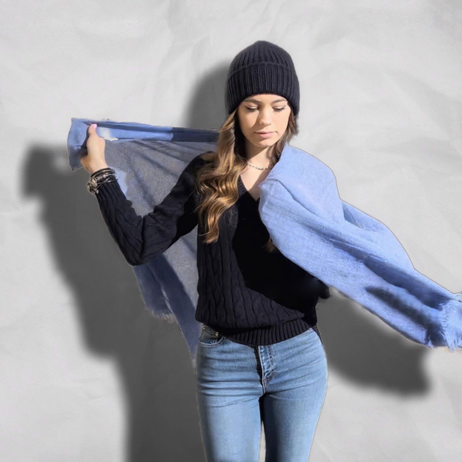 Persona con berretto e maglione neri che tiene in mano una sciarpa azzurra su sfondo grigio
