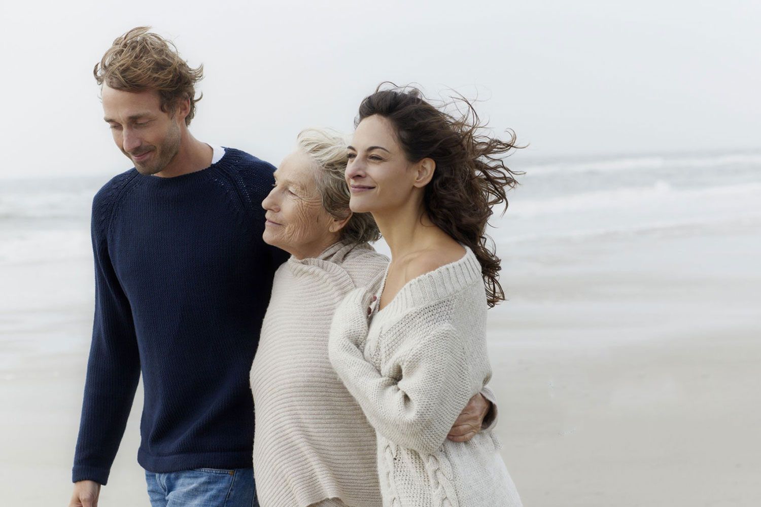 Tre persone che camminano insieme su una spiaggia nebbiosa, sorridenti e con indosso dei maglioni leggeri.