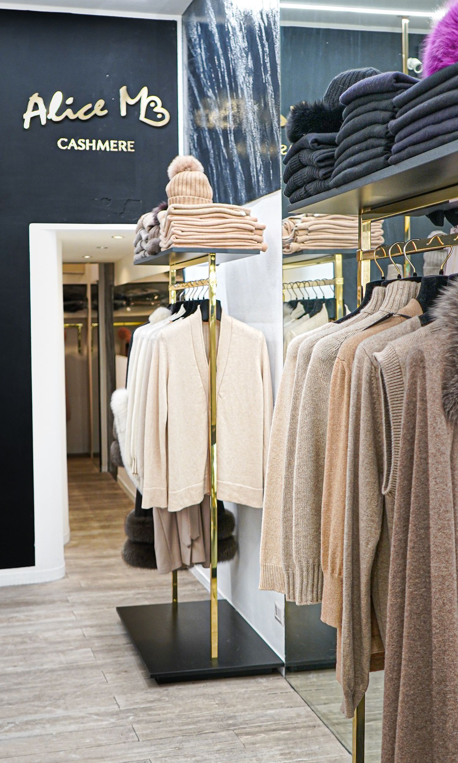 Interno di una boutique di abbigliamento in cashmere con maglioni color crema appesi a delle grucce e un'insegna nera a muro con la scritta "Alice MB".
