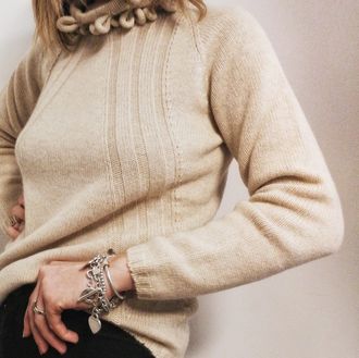 Primo piano di un maglione beige lavorato a maglia con scollo intrecciato e braccialetti d'argento indossato da una persona seduta.