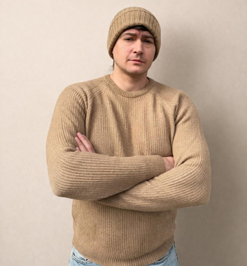Persona con maglione e berretto di lana beige, in piedi con le braccia incrociate contro un muro spoglio.