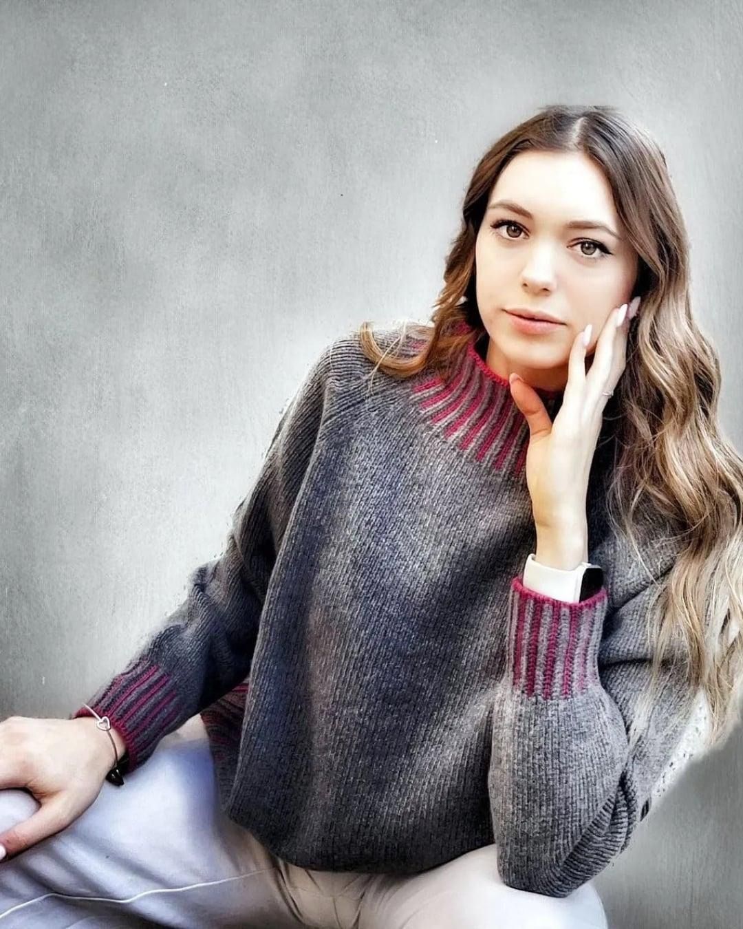 Una donna con un maglione grigio dalle maniche color bordeaux è sdraiata contro una parete spoglia con la mano sulla guancia.