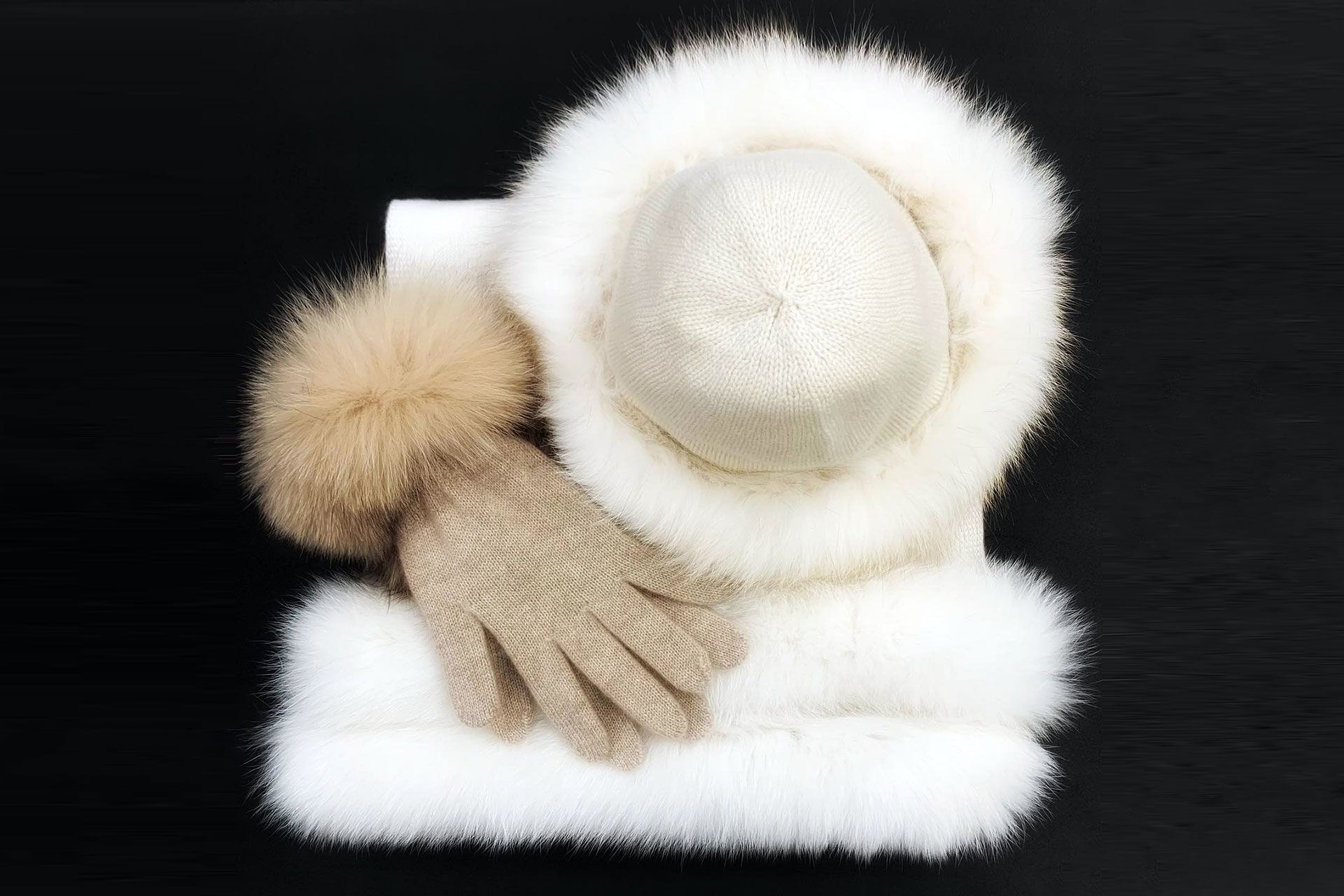 Cappello e sciarpa di pelliccia bianca con guanti beige e pon-pon, disposti su sfondo nero.