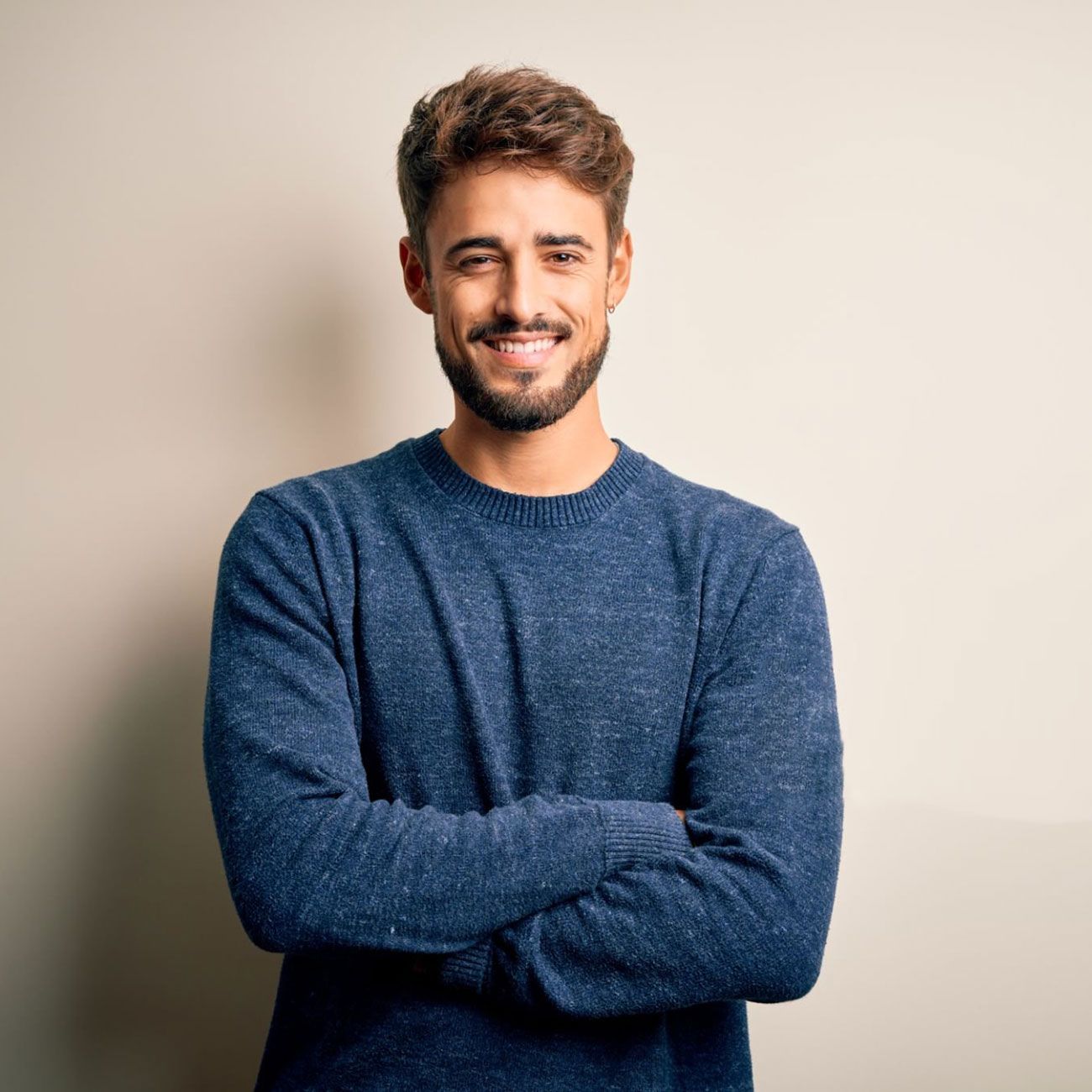 Uomo sorridente con maglione blu e braccia incrociate su sfondo beige