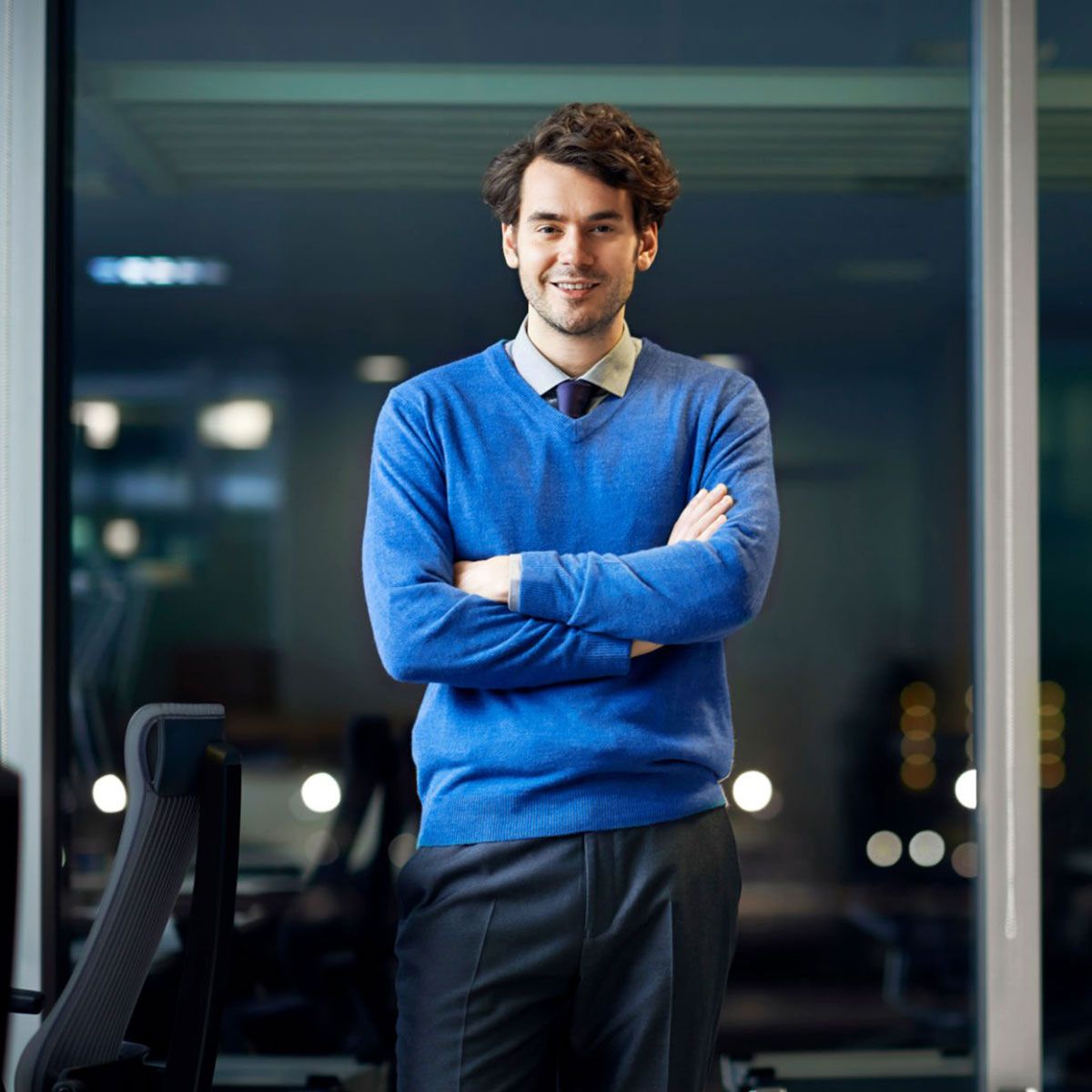 Uomo con maglione blu in piedi con le braccia incrociate in un ufficio moderno di notte
