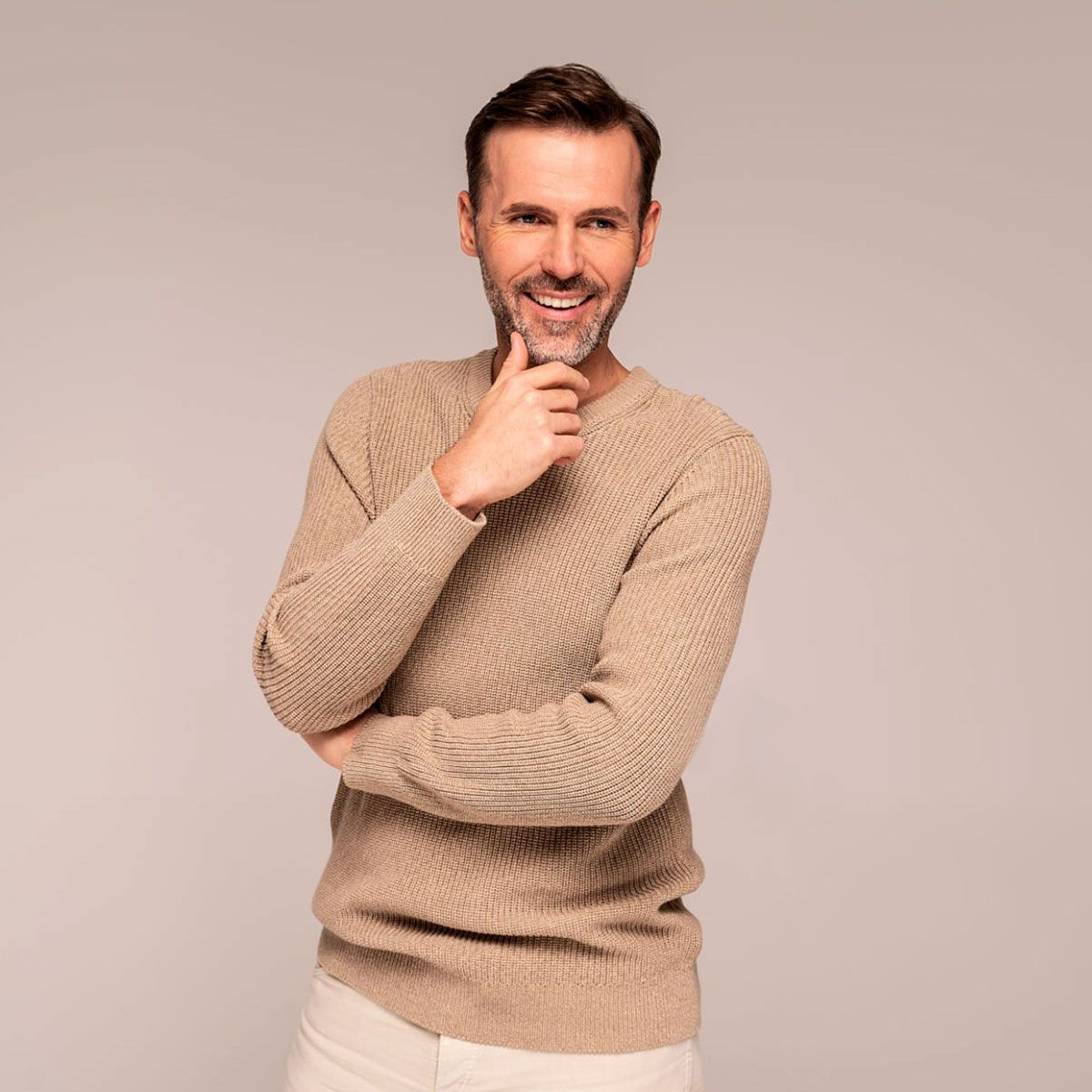 Uomo sorridente con maglione beige, braccia incrociate e mano sul mento, su sfondo neutro.