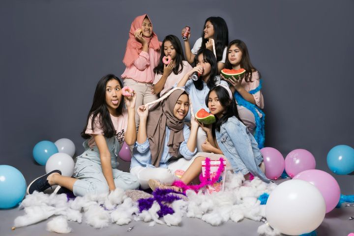 Group posing on a fluffy white set with pastel balloons, smiling and holding colorful props against a gray backdrop