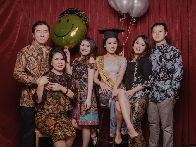 Seven people posing at a graduation celebration with balloons and a red curtain backdrop.