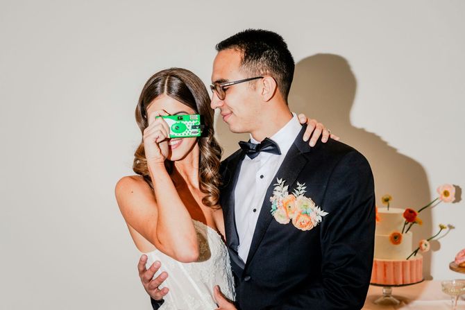 Bride and groom posing at a wedding, she covers her face with a green camera while he smiles beside her
