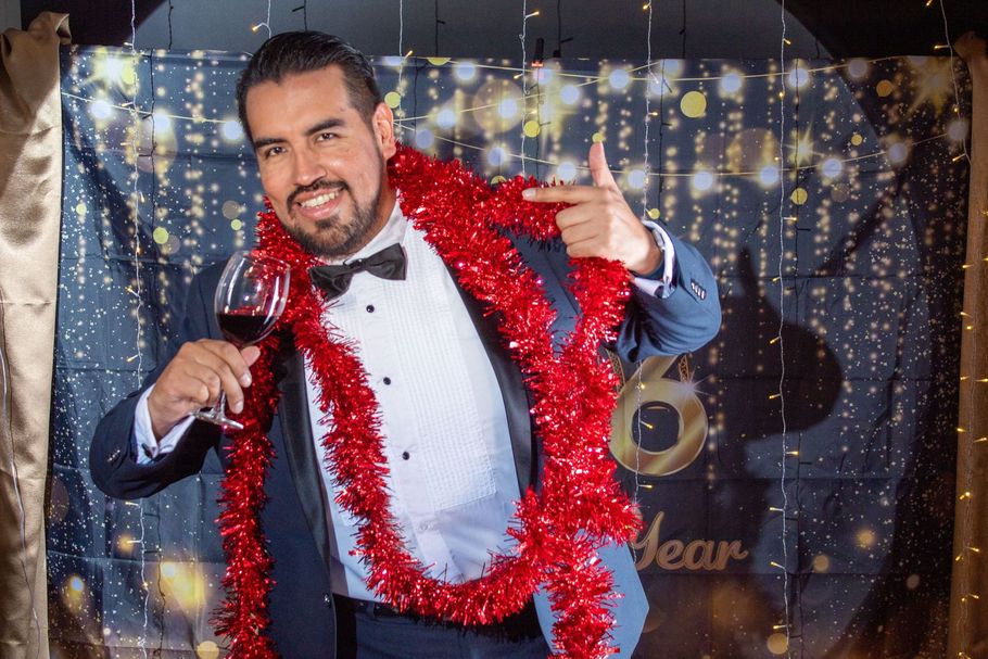 Man smiling with a wine glass and red tinsel scarf at a festive party backdrop