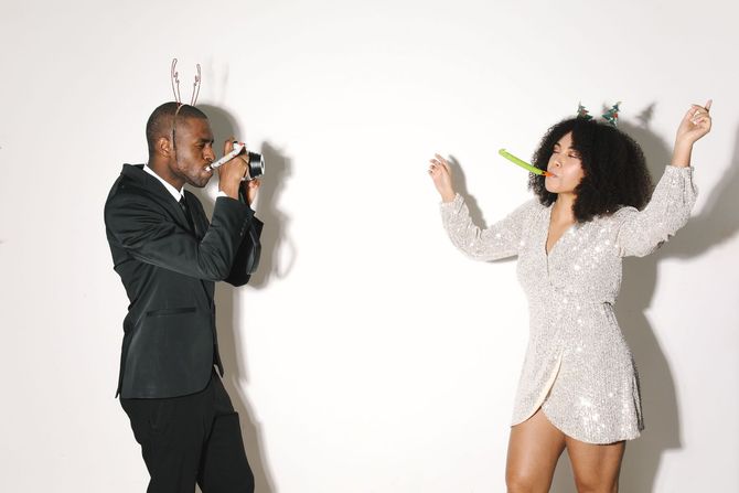 Two people dancing against a white wall, one holding a camera-like object, the other with arms raised.
