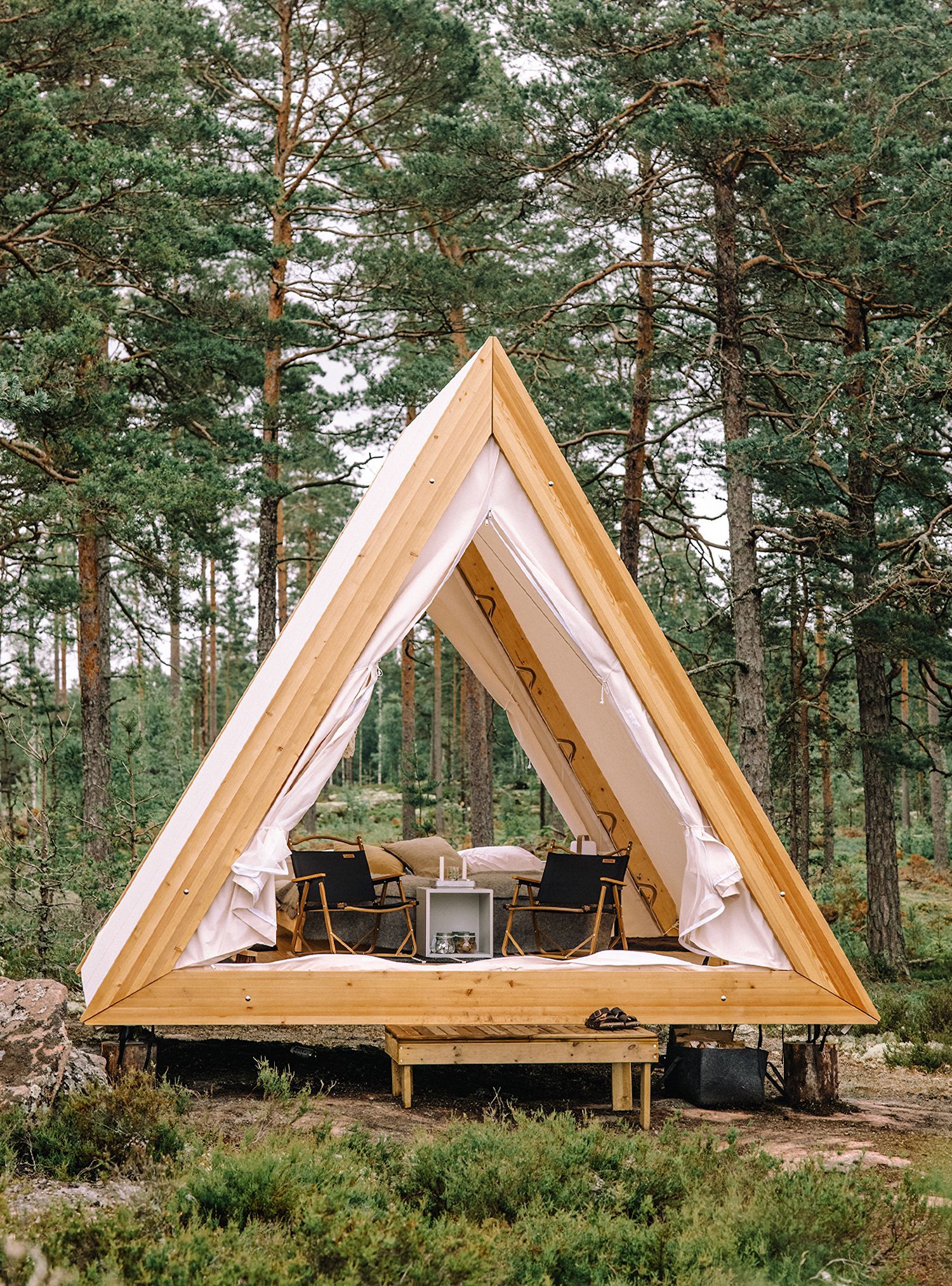 Triangle-shaped cabin with wooden frame, canvas walls, and platform. Set in a forest with chairs and table inside.