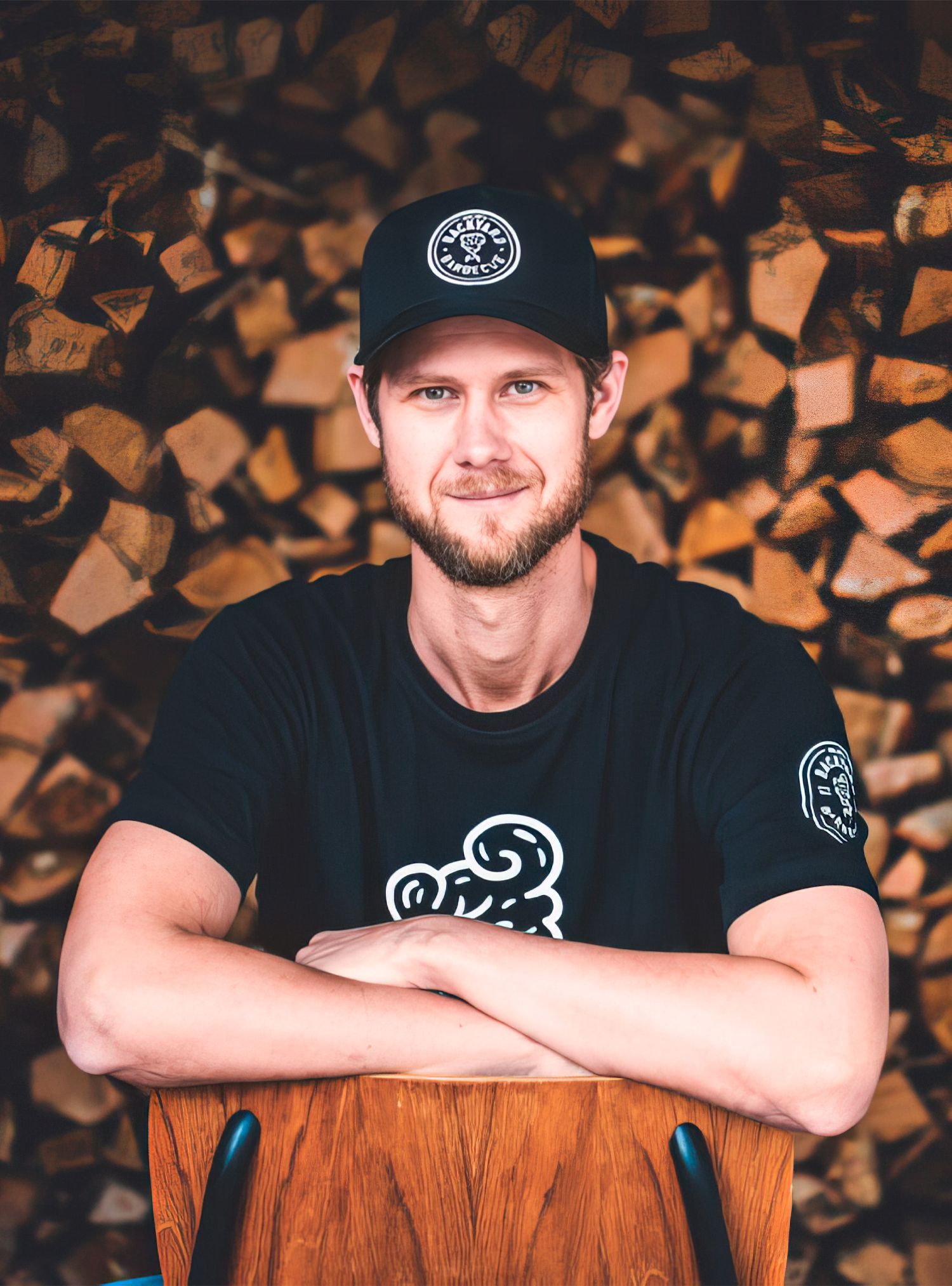 Man in black shirt and cap smiles, arms crossed, seated on a wooden chair, with a backdrop of firewood.