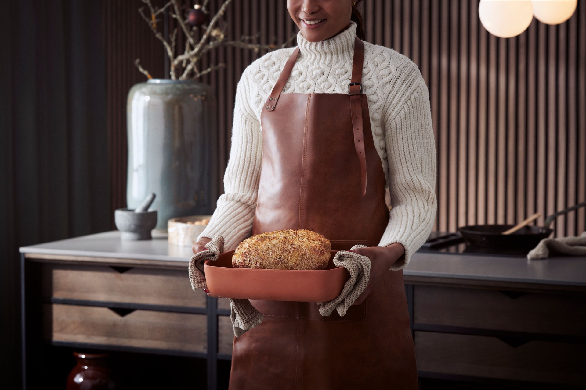Woman holding a baking pan with bread, wearing a leather apron and sweater in a kitchen.