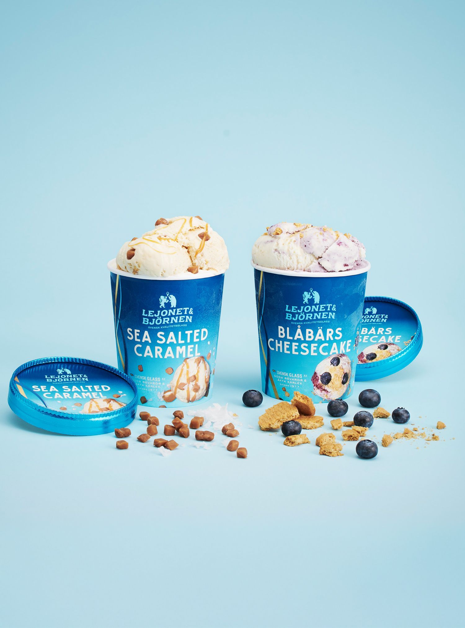 Two ice cream cups: Sea Salted Caramel and Blueberry Cheesecake, with toppings, lids, and ingredients on blue.