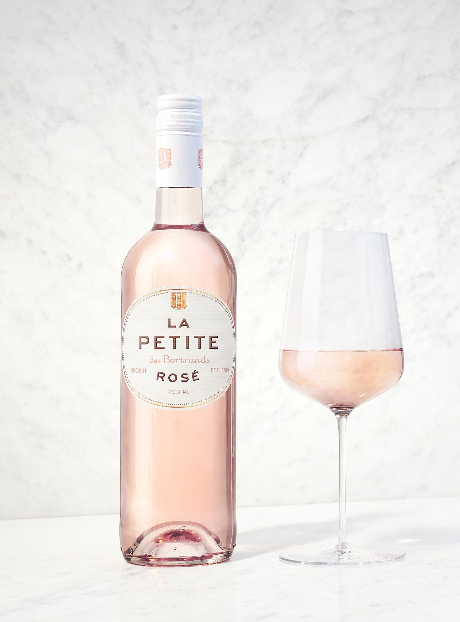 Bottle of La Petite Rosé and a glass filled with pink wine on a white surface.